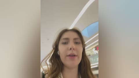 Media: A video of a young woman with wavy brown hair, fair skin, and a neutral expression. She stands in a modern, brightly lit indoor space with white walls and a curved ceiling.