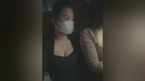 Video of two Asian women wearing face masks, seated in a dimly-lit room, one in a black dress, the other in a beige top.