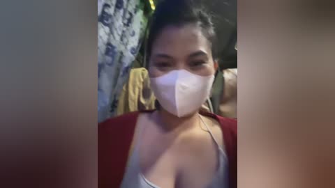 Media: Video of an Asian woman with medium skin tone and dark hair, wearing a white mask, red top, and grey tank top, standing in a dimly lit room with patterned curtains and a towel.