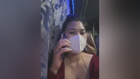 Media: Video of a woman with medium skin tone, wearing a white mask, red top, and headphones, talking on a phone, standing in a dimly lit room with blue light accents.