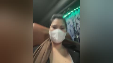 Media: Video of a woman wearing a white surgical mask and brown top, seated on a plane with green lighting, blurred background.