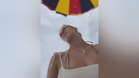 Media: Video of a woman in a beige top, leaning back with eyes closed, under a colorful umbrella, in a blurred beach background.