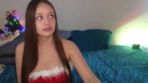 Media: Video of a young, fair-skinned woman with long, straight brown hair, wearing a red and white Santa-themed strapless dress, sitting on a blue, textured blanket in a dimly lit bedroom with a Christmas tree and multicolored lights in the background.