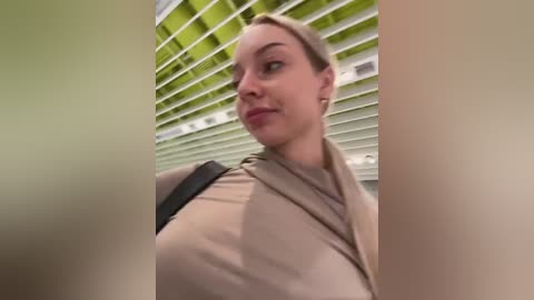 Media: Video of a smiling, fair-skinned woman with blonde hair, wearing a beige top, standing in front of green and white blinds, taken from a low angle.