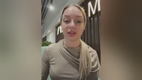 Media: Video of a young, fair-skinned woman with long blonde hair in a ponytail, wearing a beige, long-sleeve top, seated indoors with a modern, minimalist decor featuring large \"M\" letters.