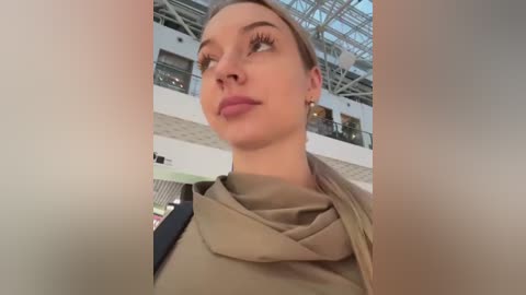 Media: Video of a fair-skinned, slim woman with light brown hair, wearing a beige scarf and light makeup, standing in a modern, glass-roofed atrium with metal beams and glass panels.