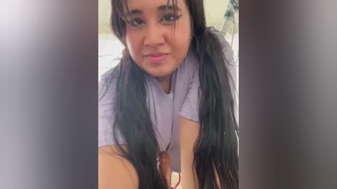 Media: Video of a young woman with long, wet, black hair, wearing a lavender shirt, leaning forward. Background is blurred, suggesting a bathroom.