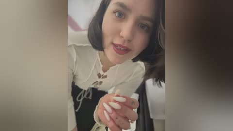 Media: A close-up video of a young woman with pale skin, dark hair, and light makeup, wearing a white lace-up blouse and black skirt, taken from a high angle.