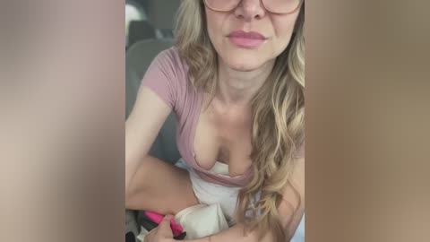 Media: Video of a fair-skinned woman with long, wavy blonde hair, wearing glasses and a low-cut pink top, sitting in a car, holding a pink cell phone.