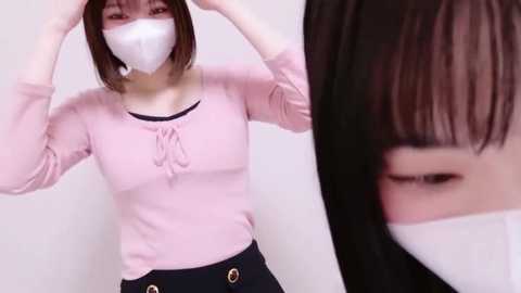 Media: Video of a woman with short brown hair, wearing a pink long-sleeved top, black skirt, white mask, adjusting her mask.