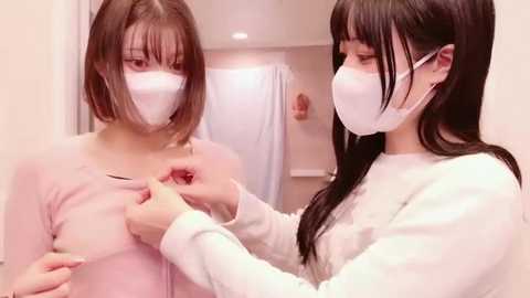 Media: Video of two women with short hair and white masks in a bathroom, one applying a red liquid to the other's chest.