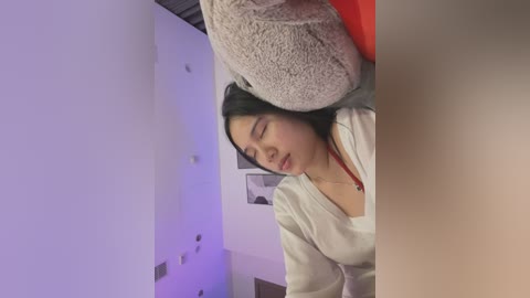 Media: Video of a young Asian woman with long black hair, wearing a white top, lying on a bed with a beige pillow, surrounded by white walls and a red blanket.