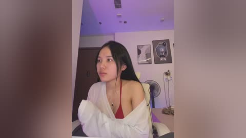 Media: A video of an Asian woman with long black hair, wearing a white off-shoulder sweater over a red top, seated indoors with a fan and framed art in the background.