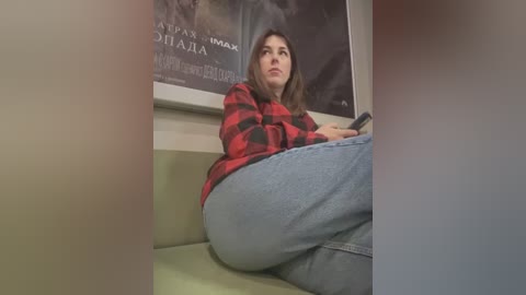 Media: A video of a young woman with light skin and long brown hair, wearing a red and black flannel shirt and blue jeans, sitting on a green sofa in a dimly lit room.