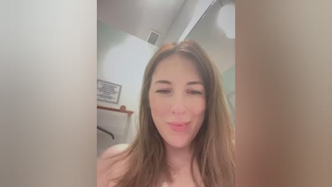 Media: Video of a fair-skinned woman with long brown hair, smiling, in a bathroom with beige walls, a wooden railing, and a partially visible mirror.