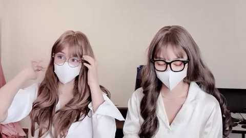 Media: Video of two women with long brown hair, wearing white lab coats and face masks, adjusting their glasses, sitting indoors.