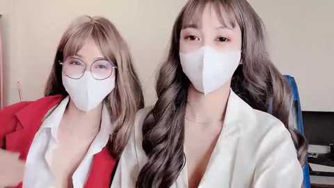 Media: Video of two East Asian women with long, wavy brown hair, wearing white face masks, red blazers, and glasses. One has a deep cleavage, sitting in a blue chair, while the other wears a white blouse.