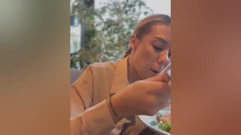 Media: A video of a young woman with light skin, brown hair, wearing a beige jacket, intently drinking from a cup, with a blurred background of greenery and a building.