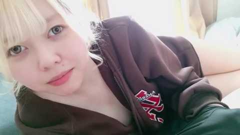 Media: Video of a young Asian woman with fair skin and blonde hair, wearing a dark brown hoodie with red and white insignia. She is lying on a bed with light-colored sheets in a softly lit room.