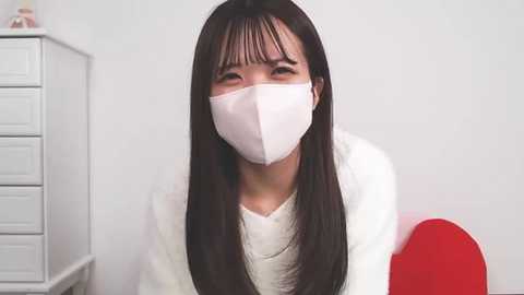 Media: Video of an East Asian woman with long black hair, wearing a white face mask, white sweater, and a white wall in the background.