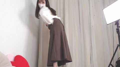 Media: A video of a slender woman with long dark hair, wearing a white turtleneck and high-waisted brown skirt, standing against a white backdrop with a studio light to the right.