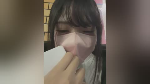 Media: Video of a young Asian woman with long black hair, wearing a pink face mask, intently reading a white paper with a hand. Background features a brick wall and a pink wall.