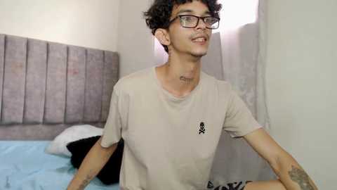 Media: Video of a young man with curly hair, wearing glasses, beige shirt, and black pants, sitting on a bed with a grey headboard, in a simple, modern bedroom.