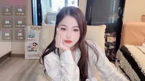 Media: Video of an Asian woman with long black hair, fair skin, and rosy cheeks, wearing a white plaid shirt, resting her chin on her hand. She's in a cozy, minimalist room with beige furniture, a calendar, and a window.