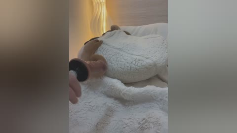 Media: Video of a plush teddy bear being held by a hand with a black ring. The bear is white, fuzzy, and has a textured, quilted look. The background is soft and dimly lit, with a warm, ambient glow.