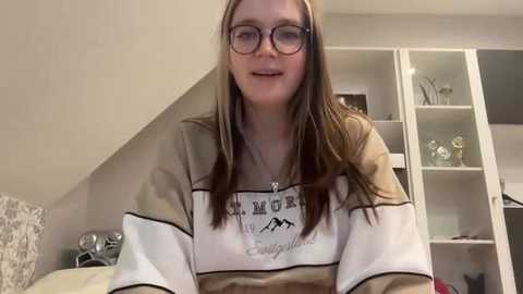 Media: Video of a young woman with fair skin, long brown hair, and glasses, wearing a beige and white striped hoodie. She is indoors, in a room with a white bookshelf and a bed with a patterned pillow.