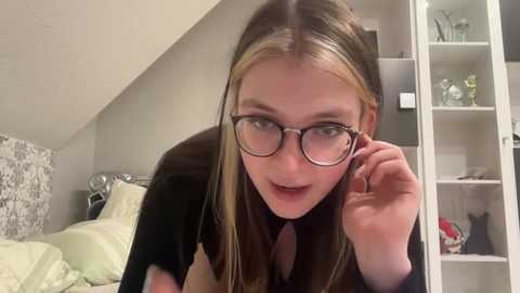 Media: Video of a young Caucasian woman with straight, shoulder-length blonde hair, wearing glasses, and a black top, leaning on a bed in a modern, well-lit bedroom.