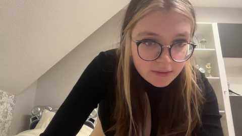 Media: A video of a young woman with long, light brown hair, wearing black-framed glasses and a black sweater, leaning forward in a bedroom with a white bed and patterned wall.