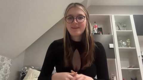 Media: Video of a young woman with long, straight brown hair, wearing glasses, a black top with a deep V-neck, and a slight smile. Background includes a modern, minimalistic bedroom with white furniture and a patterned pillow.