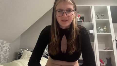 Media: Video of a young woman with fair skin, long brown hair, wearing glasses and a black long-sleeve crop top with a cutout revealing cleavage. She stands in a modern bedroom with a white wardrobe and patterned bedspread.