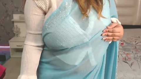 Media: Video of a woman with light skin, wearing a light blue sari with glittery details, holding a hand to her chest. She has long, straight, blonde hair. Background includes a gray and floral-patterned wall, a white nightstand, and a pink object.