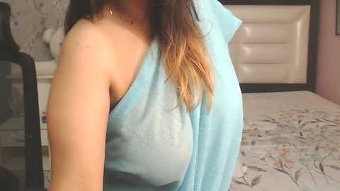 Media: A video of a woman with light skin and dark brown hair, wearing a light blue, sleeveless top, sitting in a bedroom with white bedding, floral design, and a white headboard.