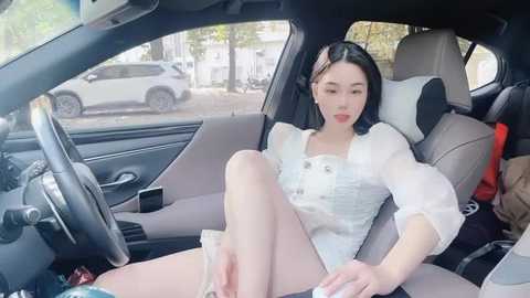 Media: Video of an East Asian woman with fair skin and long black hair, wearing a white blouse and white shorts, sitting seductively in a car, with a smartphone in hand.