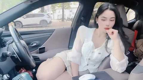 Media: Video of an Asian woman with fair skin, long black hair, and makeup, sitting in a car. She wears a white, sheer blouse and shorts, looking thoughtful, with a red bag beside her.