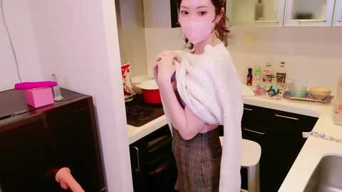 Media: Video of an Asian woman in a modern kitchen, wearing a mask, white shirt, and plaid skirt, holding a pink bucket. Background includes a stove, cabinets, and countertop cluttered with utensils.