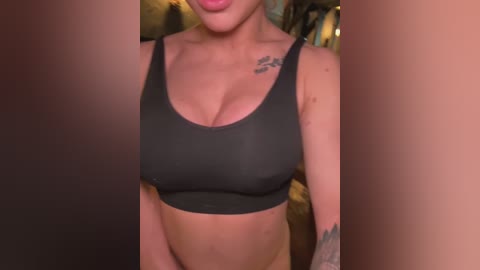 Media: A video of a light-skinned woman wearing a black sports bra, showing off her medium-sized breasts and tattoos on her shoulders. Background features blurred, warm-toned interior.