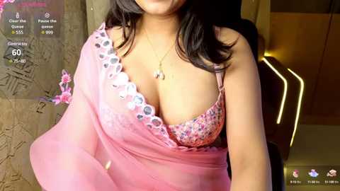 Media: A video of a South Asian woman with medium skin tone and long black hair, wearing a pink floral bra with lace detailing, and a matching sheer pink saree. The background shows a dimly lit room with a modern, minimalist decor.