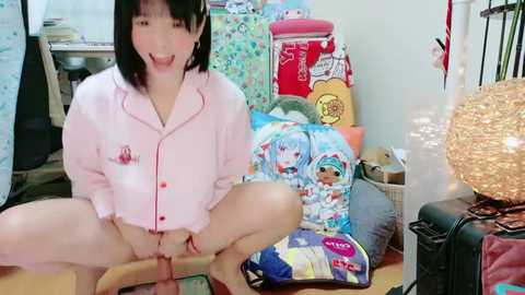 Media: A video of an Asian woman with short black hair, wearing a pink pajama shirt, squatting on a wooden floor, holding a large dildo, surrounded by colorful, cartoon-themed pillows and suitcases in a cluttered room.