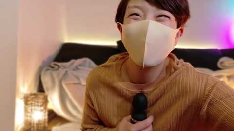 Media: Video of an Asian woman with short dark hair, wearing a beige mask, brown sweater, and holding a black microphone, smiling, in a cozy, softly lit bedroom with a bed, blankets, and ambient lighting.