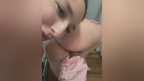 Media: A close-up video of a woman with fair skin, brown hair, and large breasts, wearing a pink bra, captured from a low angle, with blurred background of a wooden staircase.