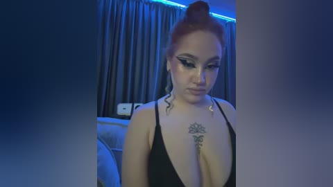 Media: Video of a fair-skinned woman with red hair in a bun, wearing black lingerie with deep V-neckline, featuring elaborate tattoos on her chest, in a dimly lit bedroom with blue lighting.