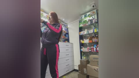 Media: Video of a person in a black and pink tracksuit, standing in a well-lit, organized pantry with white cabinets, shelves filled with food and drinks, and cardboard boxes on the floor.