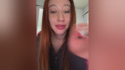 A video of a fair-skinned woman with long, straight, red hair, wearing a dark shirt, making a pouty face in front of a blurred background, possibly a bathroom.