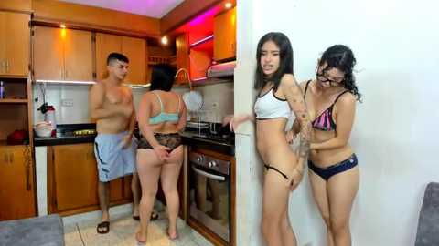 Media: Video of two Asian women in lingerie, one topless, standing beside a shirtless man in a kitchen; modern, wooden cabinetry, tiled floor, and red lighting.