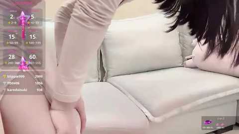 Media: Video of a young Asian woman with black hair, wearing a beige long-sleeve top, bending over a white leather couch in a minimalist room.