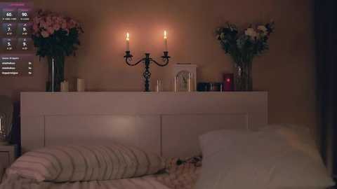 Media: Video of a dimly lit bedroom with a white bed, two lit candles on a dresser, and a bouquet of pink and white flowers on each side, creating a cozy, romantic ambiance.